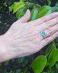 Hand wearing a natural emerald ring with accent diamonds against a leafy background.