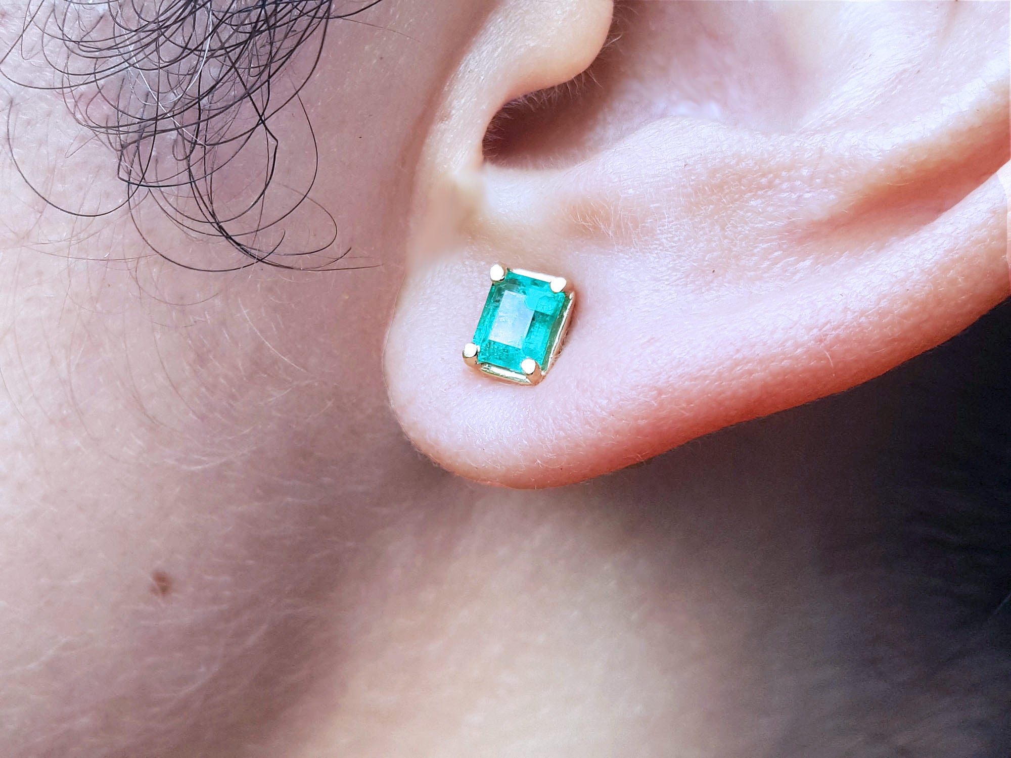 Close-up of a woman&#39;s ear wearing aemerald-cut stud emerald earring with yellow gold frame.