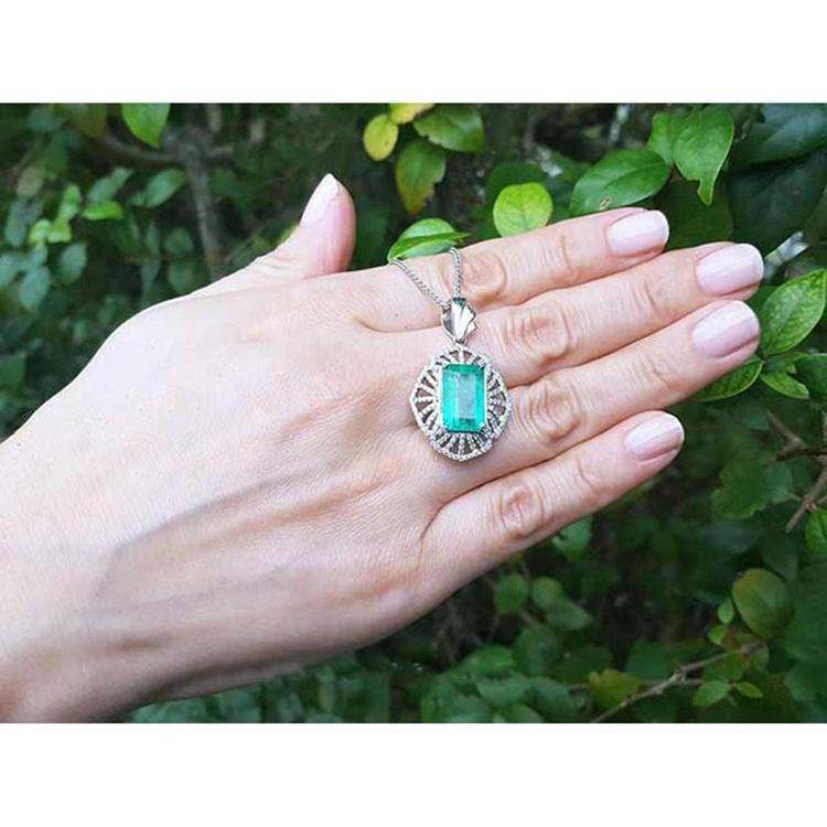 Hand holding a white gold pendant with a rectangular emerald against a green leafy background.