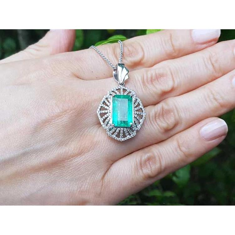 Emerald rectangular gemstone pendant on a white gold chain held in a hand with a blurred green background.
