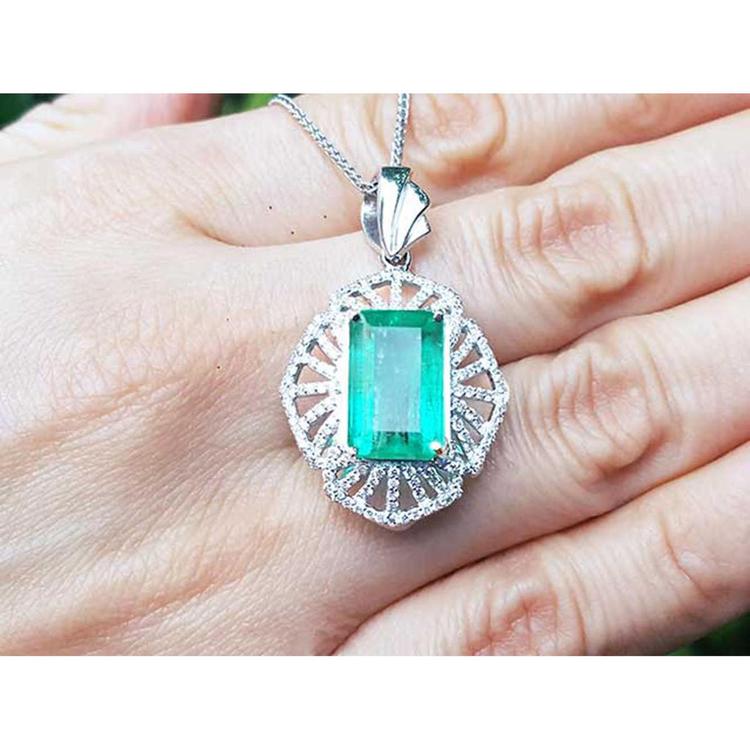 On a hand being displayed a natural emerald pendant with accent diamonds crafted in white gold.