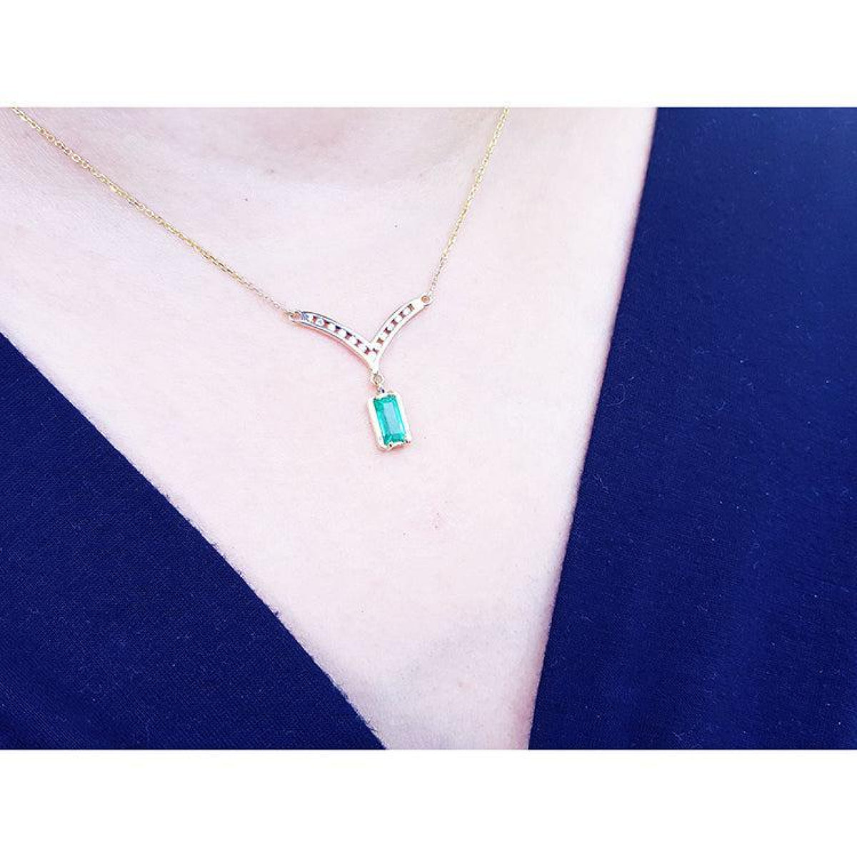 A close-up of a 14K yellow gold necklace with a baguette-cut Colombian emerald dangling from a diamond-studded V-shaped pendant, worn on a model's neck.