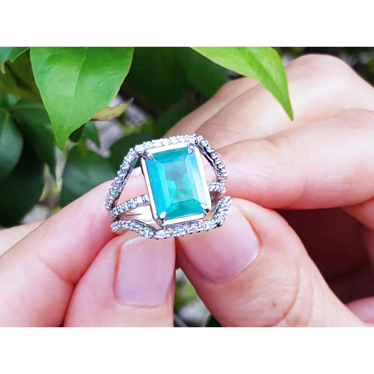 An emeral-cut-emerald ring with diamond accents held between fingers against a natural background.