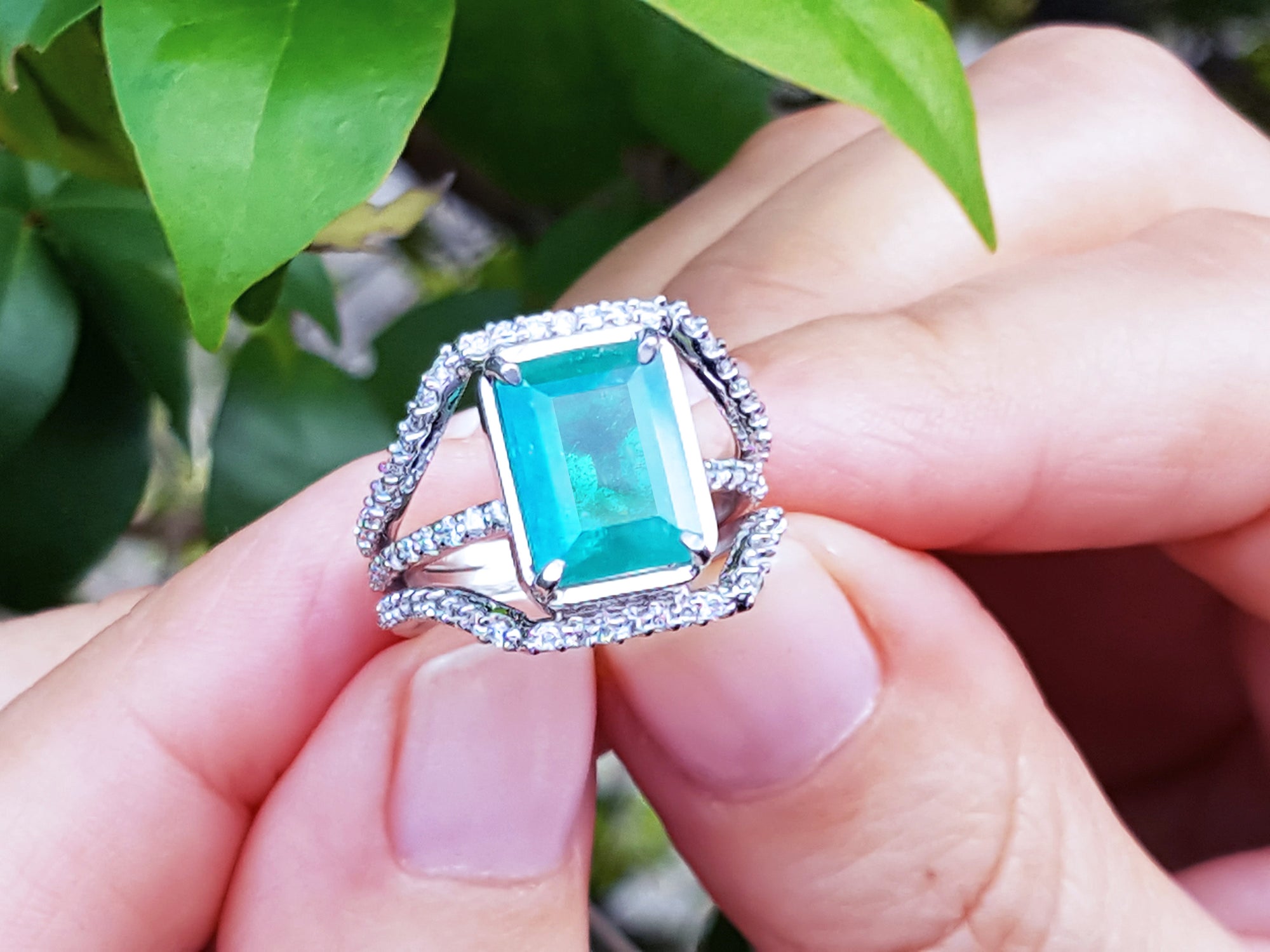 An emeral-cut-emerald ring with diamond accents held between fingers against a natural background.
