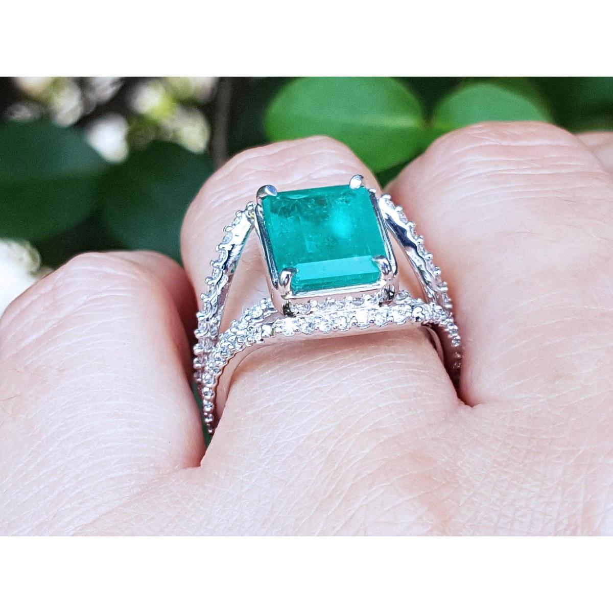 This is an emerald and diamond ring on a finger against a blurred green background.