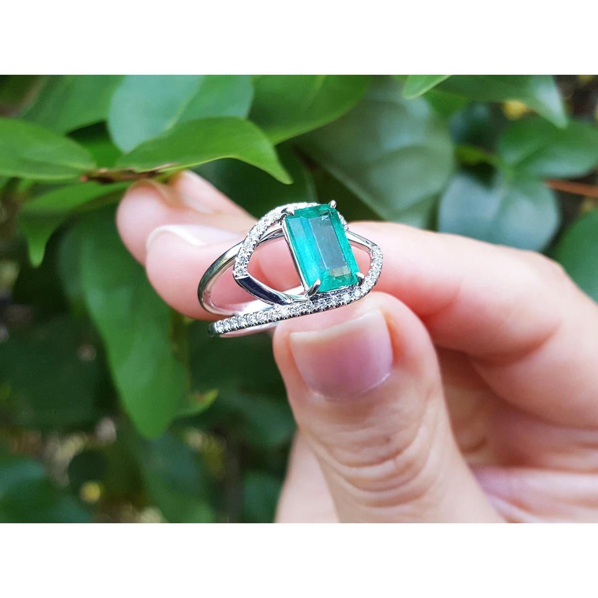 A rectangular emerald on a split shank ring held by a hand against a leafy background.