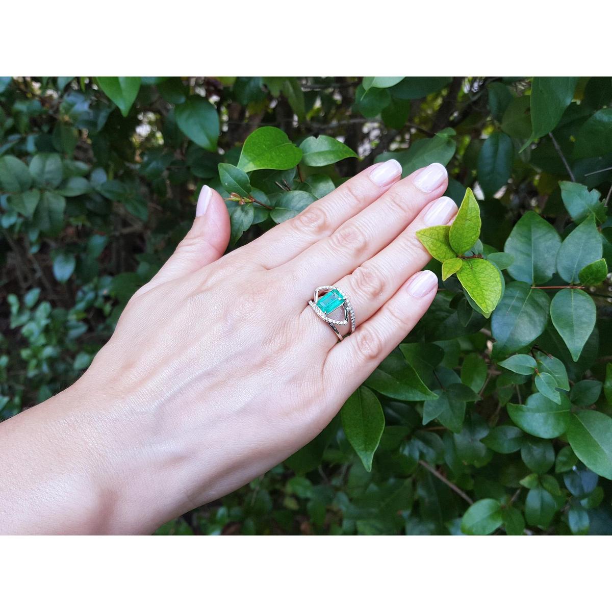 Hand wearing a white gold ring with a rectangular emerald against a green leafy background.