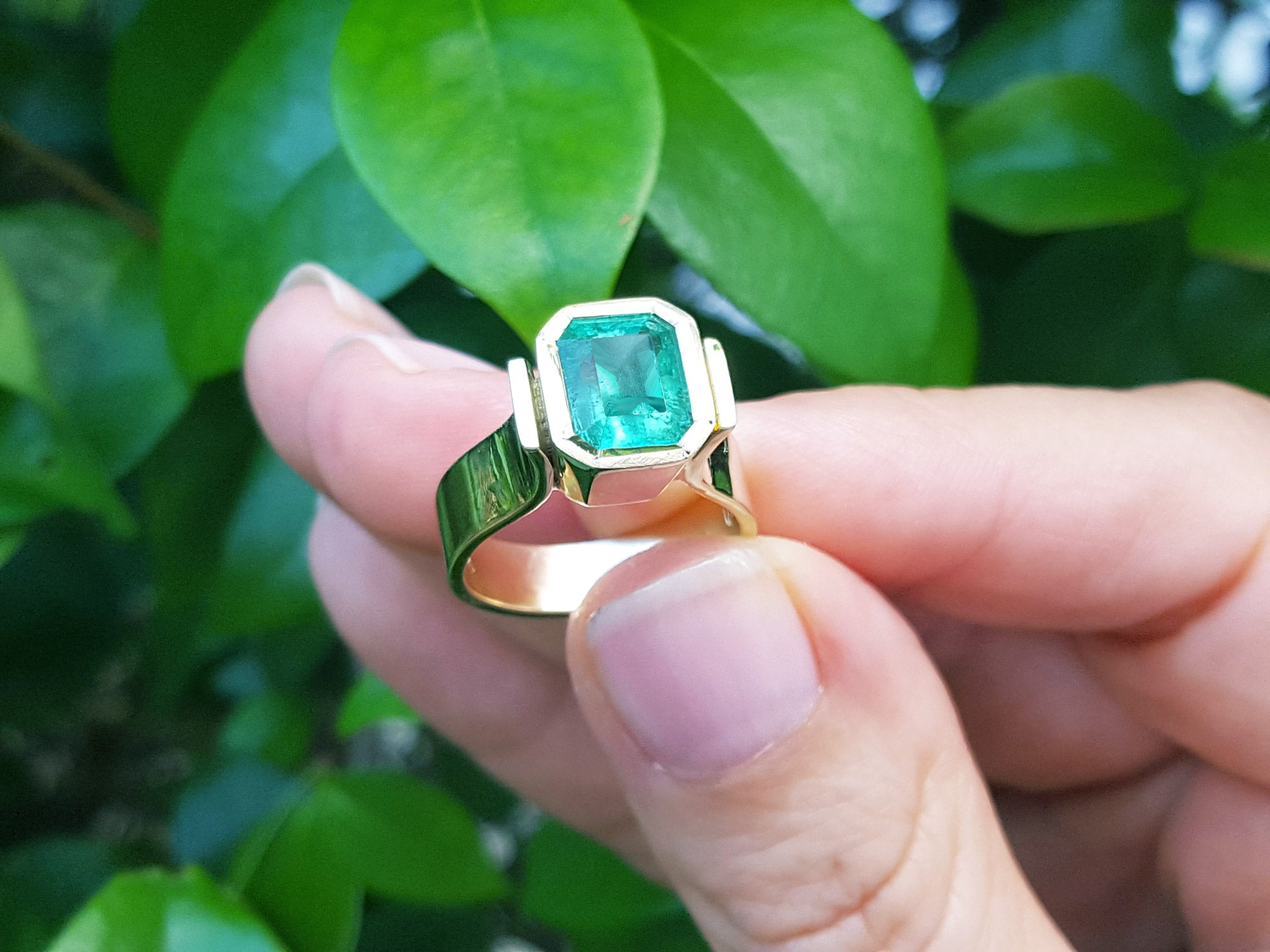 A colombian emerald solitaire ring held by a hand against a green leafy background.