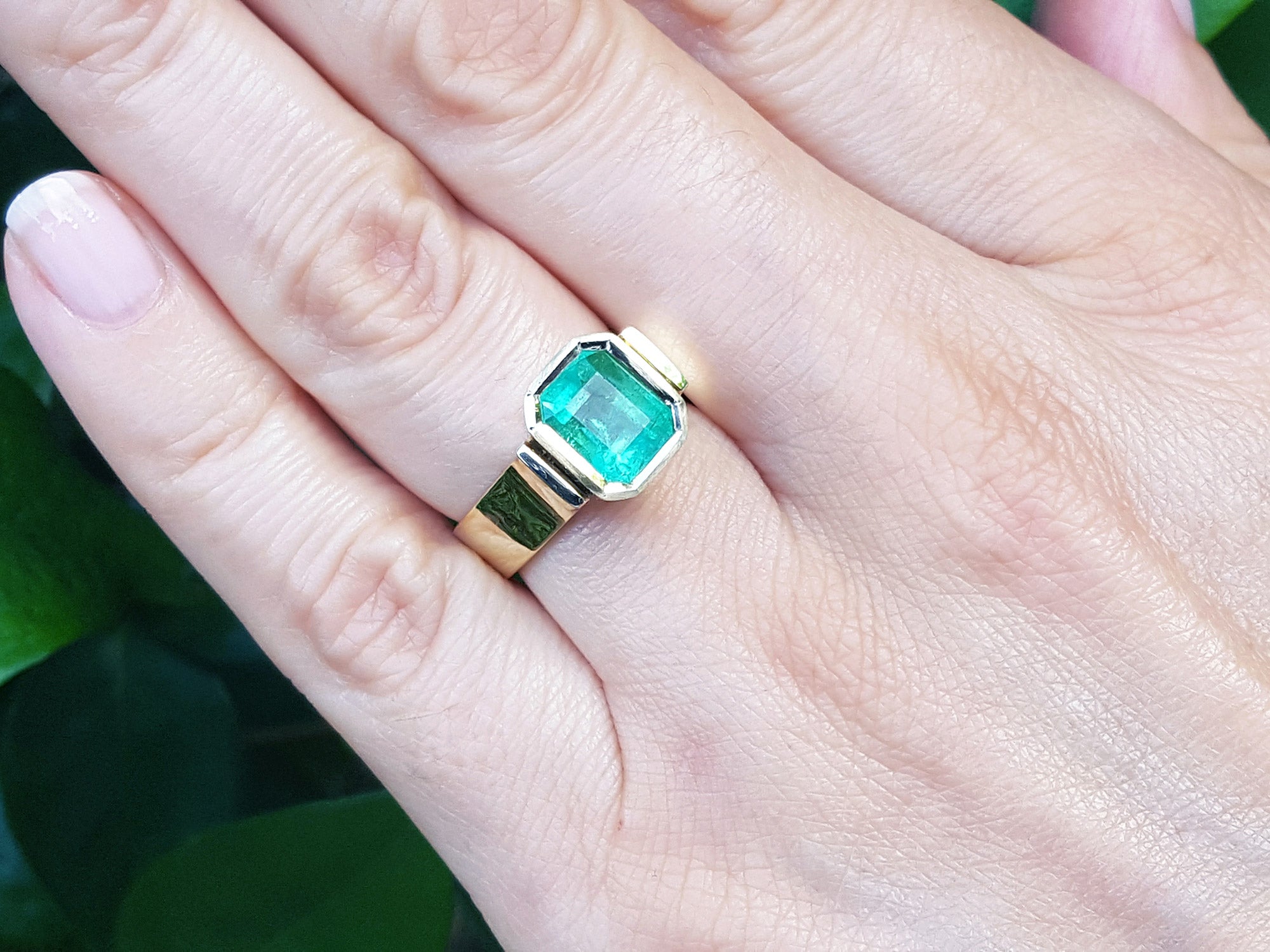A bezel set emerald solitaire ring on a person&#39;s hand against a green leafy background.