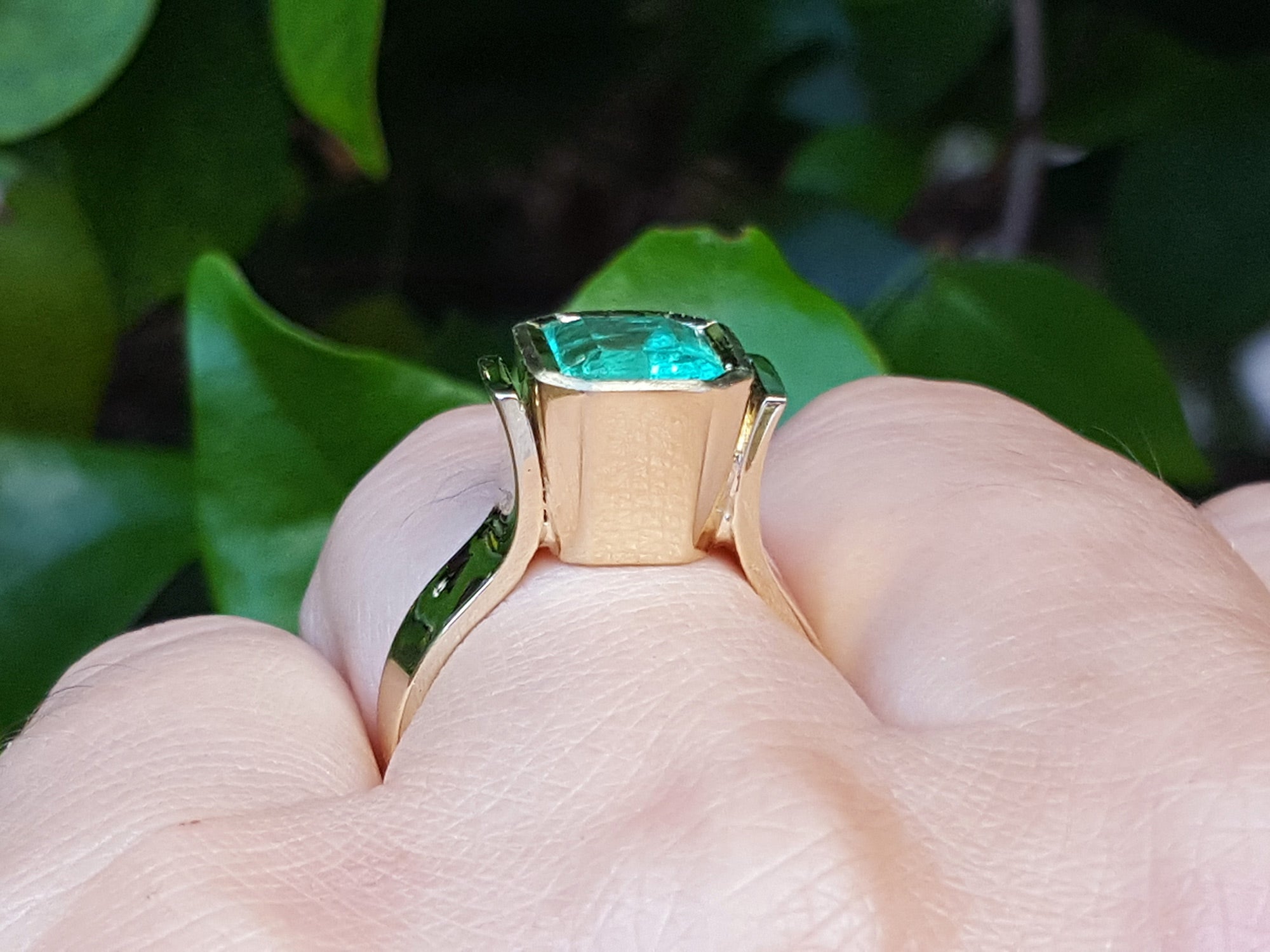 Viewing the underside of a natural emerald solitaire ring on a finger against a green leafy background.