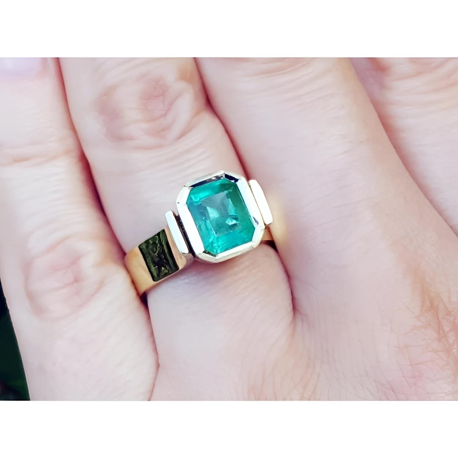 A women's solitaire emerald ring bezel set in yellow gold on a finger.