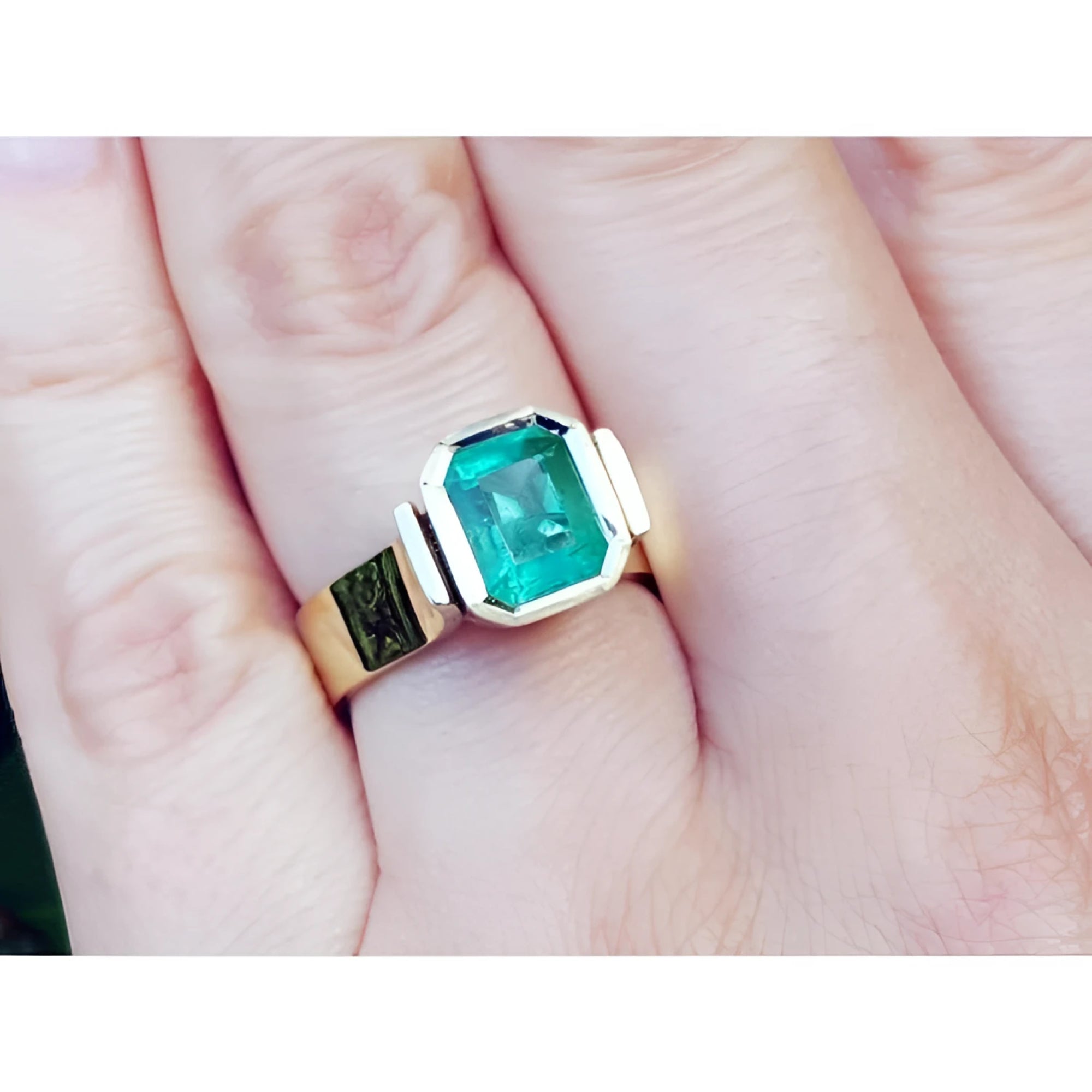 A women's solitaire emerald ring bezel set in yellow gold on a finger.