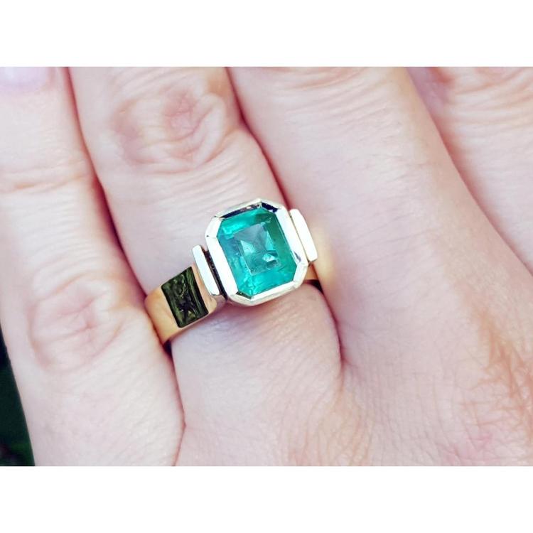 A women's solitaire emerald ring bezel set in yellow gold on a finger.