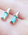 Baguette cut emerald and diamond stud earrings on a hand with a blurred green background
