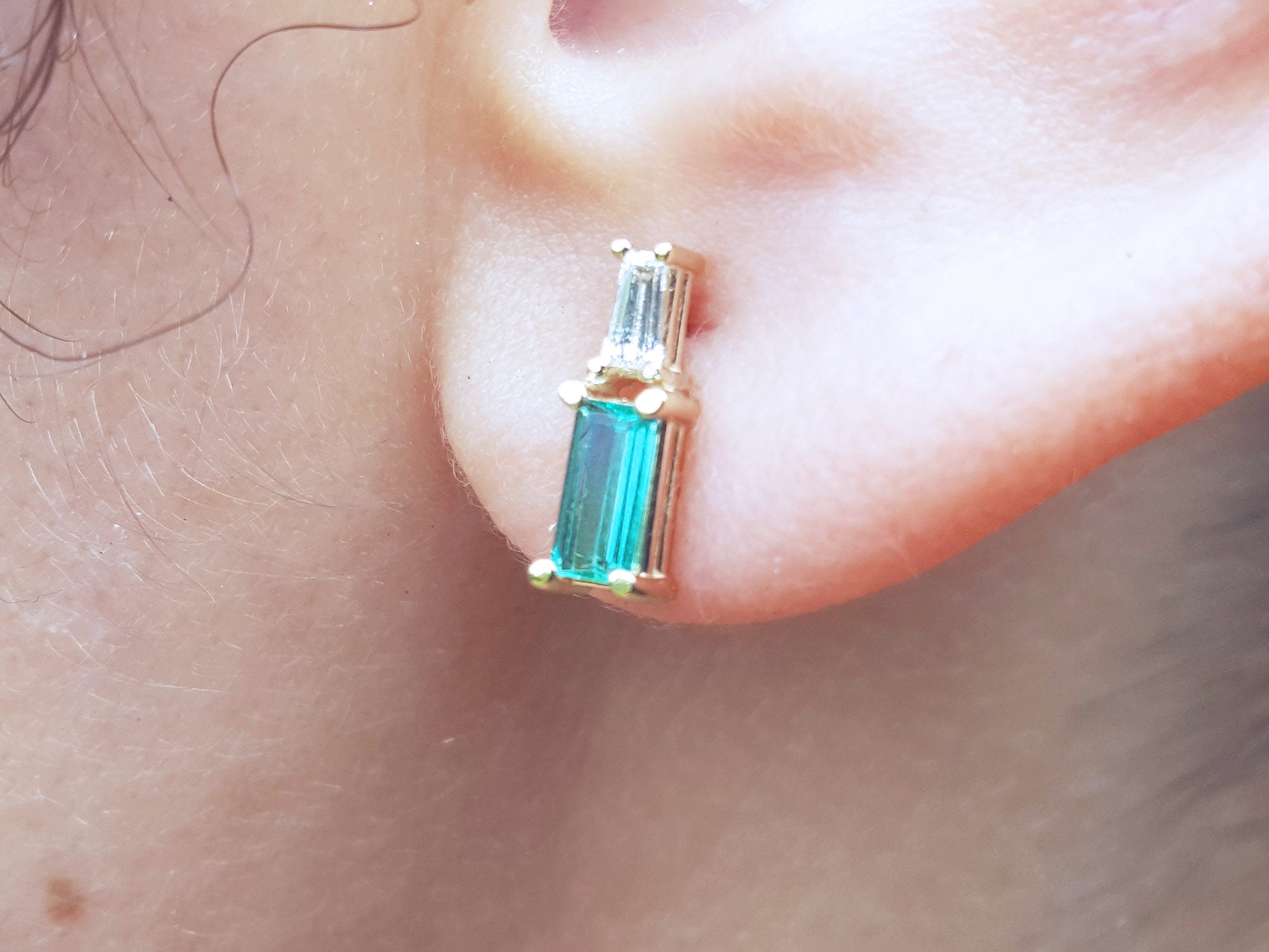Natural baguette cut emerald and diamond stud earrings worn on an ear.