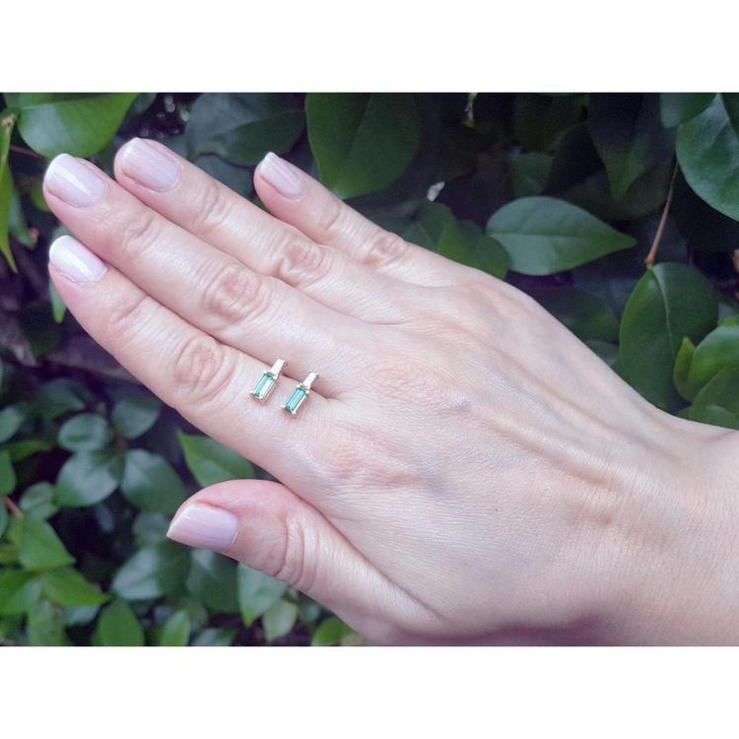 Minimalist 14K gold stud earrings with baguette emeralds and accent diamonds on a hand.