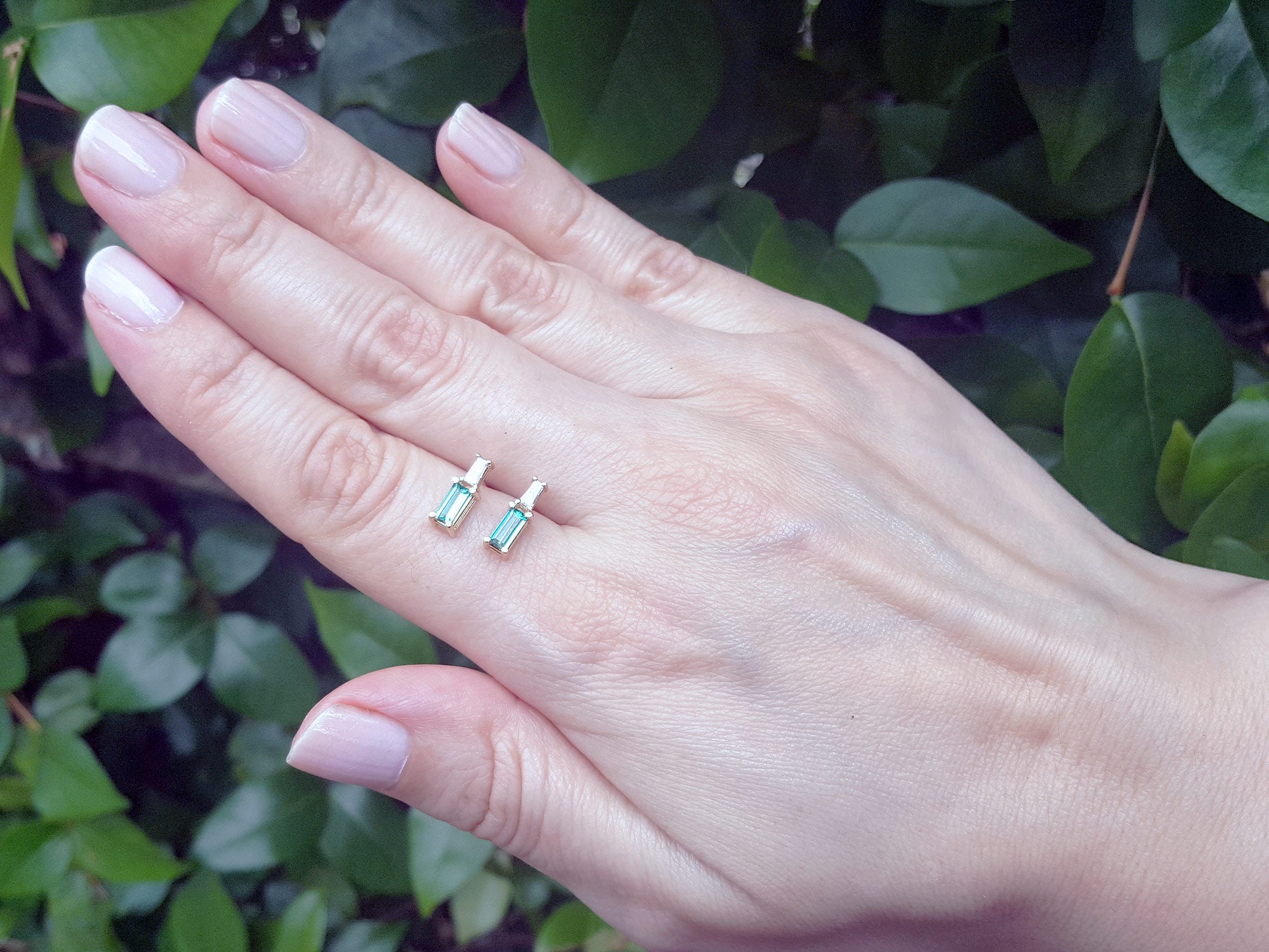 Two small emerald earrings held between fingers against a blurred green background.