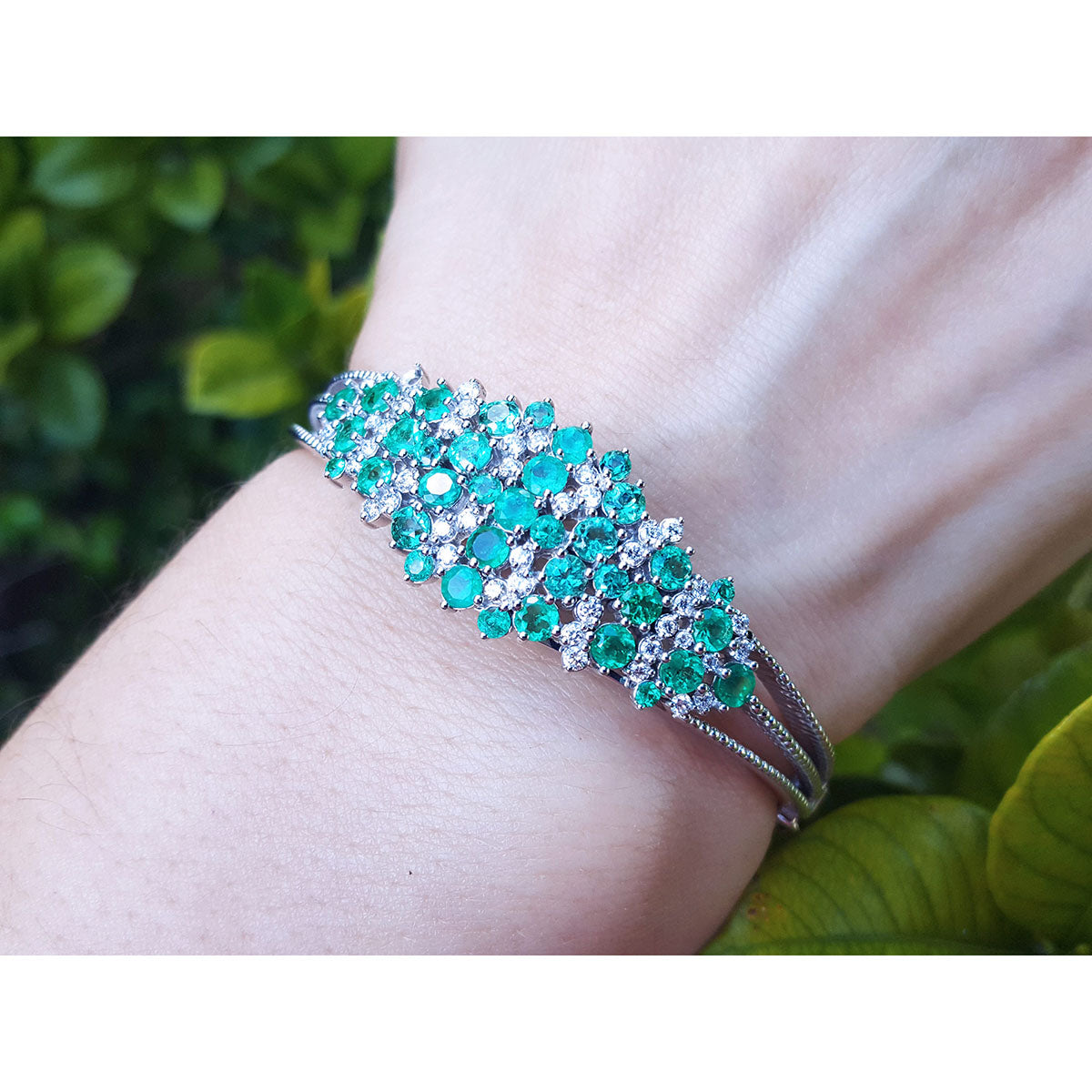 Bangle with emerald and diamonds on a wrist against a natural background.