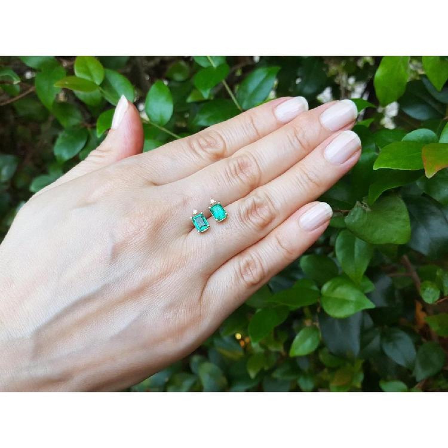14K yellow gold stud earrings with rectangular Colombian emeralds and accent diamonds, worn on a hand.