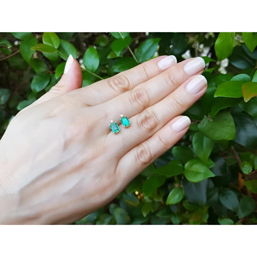 14K yellow gold stud earrings with rectangular Colombian emeralds and accent diamonds, worn on a hand.