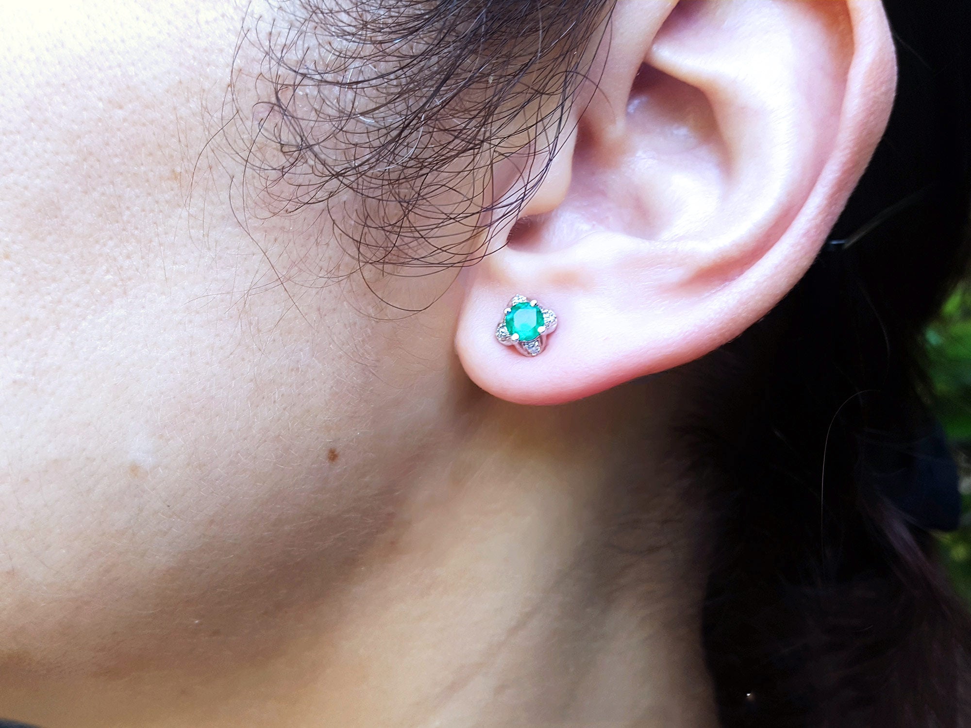 Close-up of a woman's ear wearing a tulip emerald and diamond stud earring with a blurred background