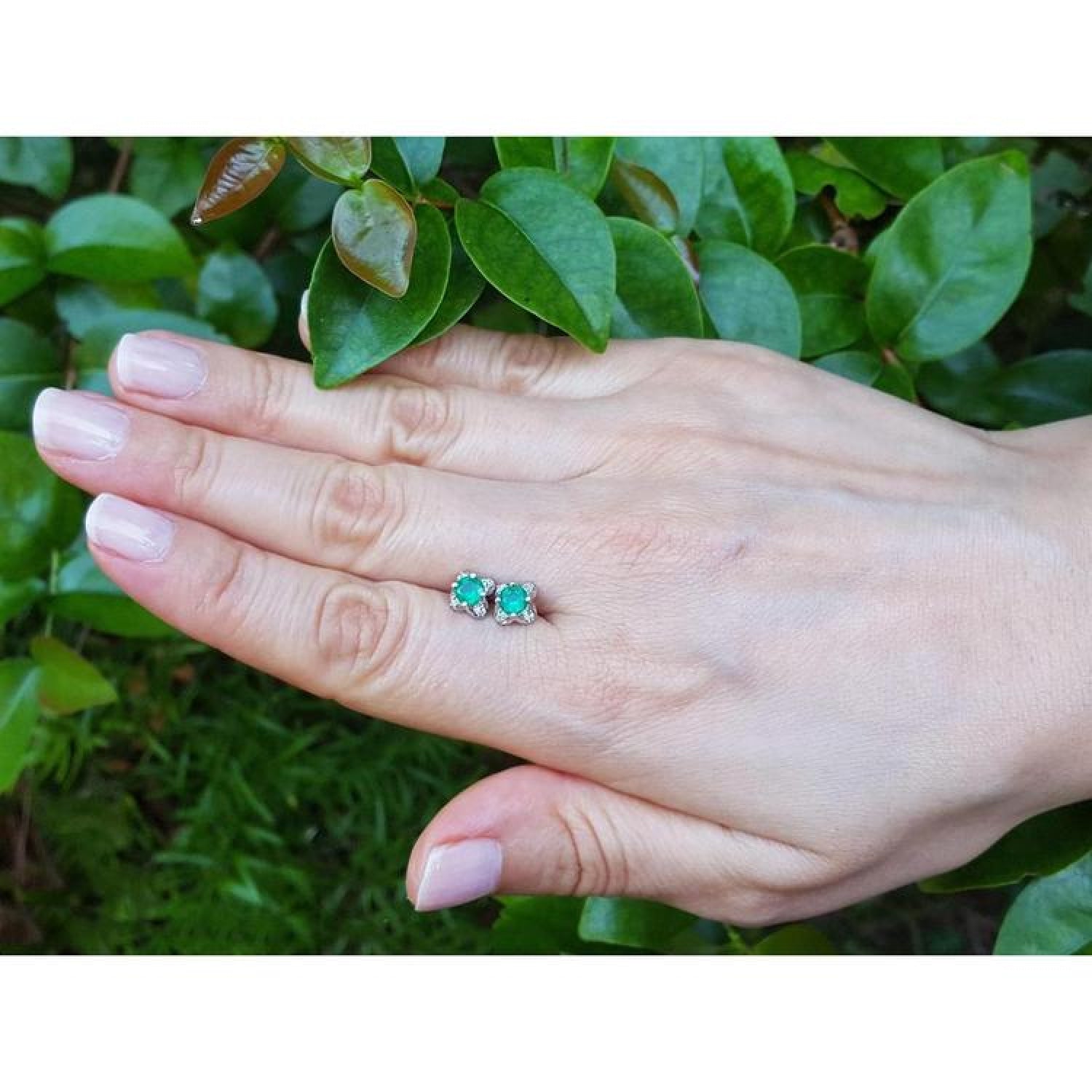 Emerald and diamond tulip stud earrings in 14K white gold, shown on a hand against green foliage.