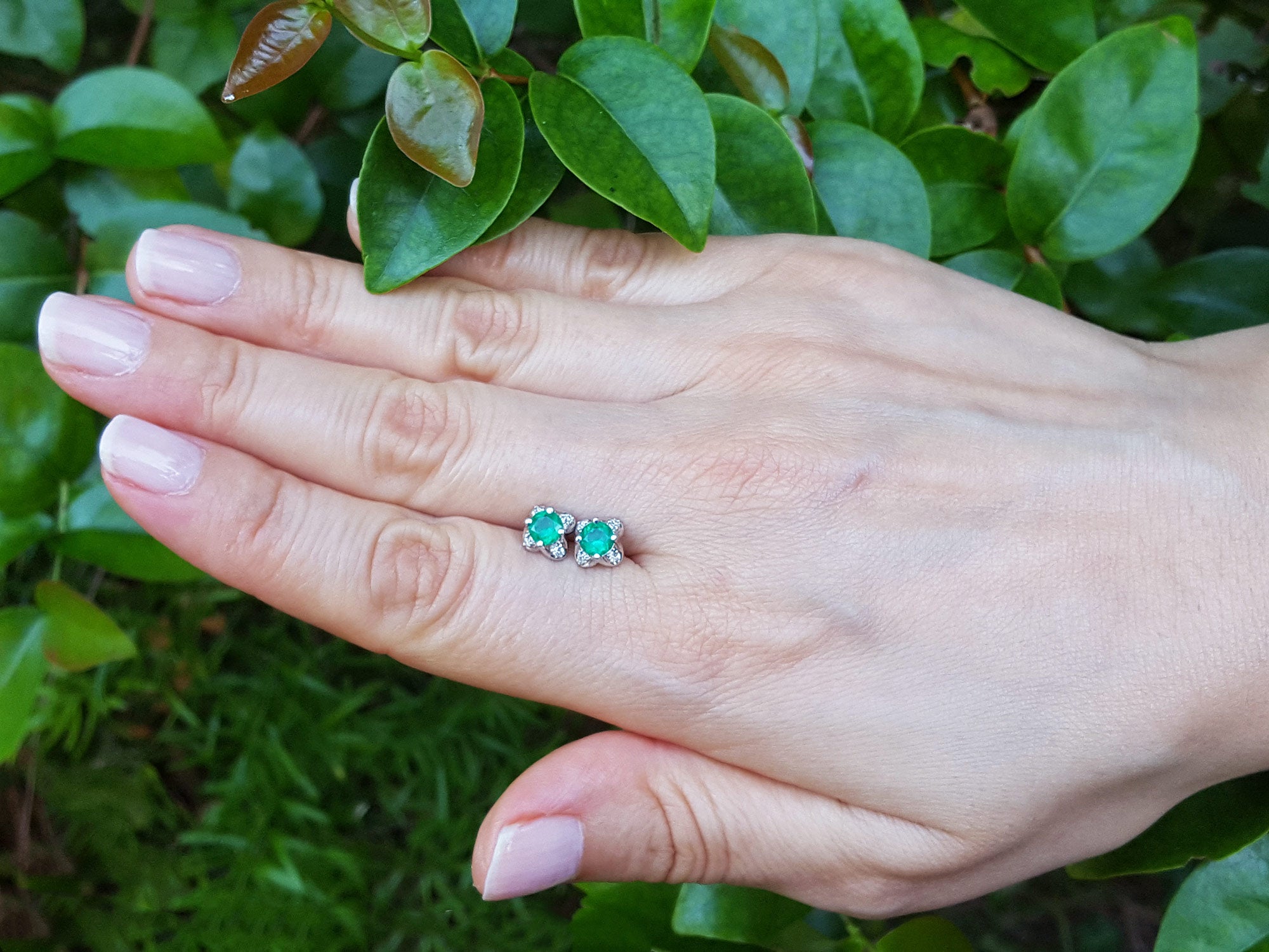 Hand holding a pair of Muzo Colombian emerald tulip stud earrings against a green leafy background.