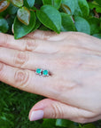 Hand holding a pair of Muzo Colombian emerald tulip stud earrings against a green leafy background.