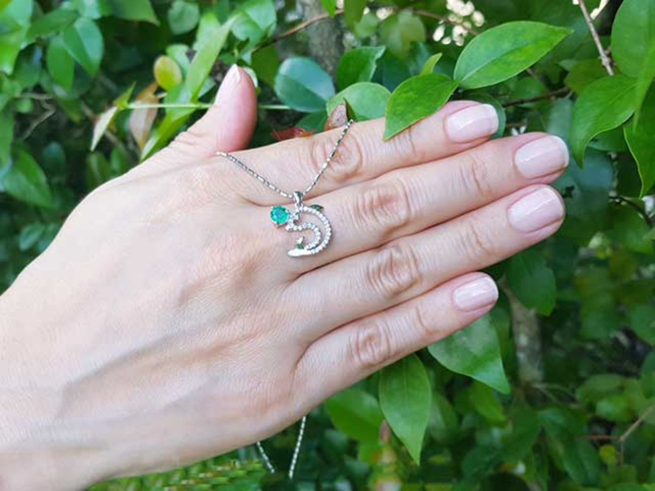 Hand displaying a round cut emerald dolphin pendant against a green leafy background.