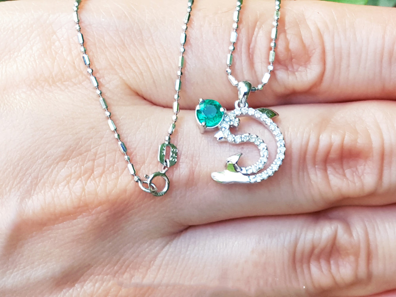 A natural round cut emerald dolphin pendant with diamonds in white gold on a person&#39;s fingers.