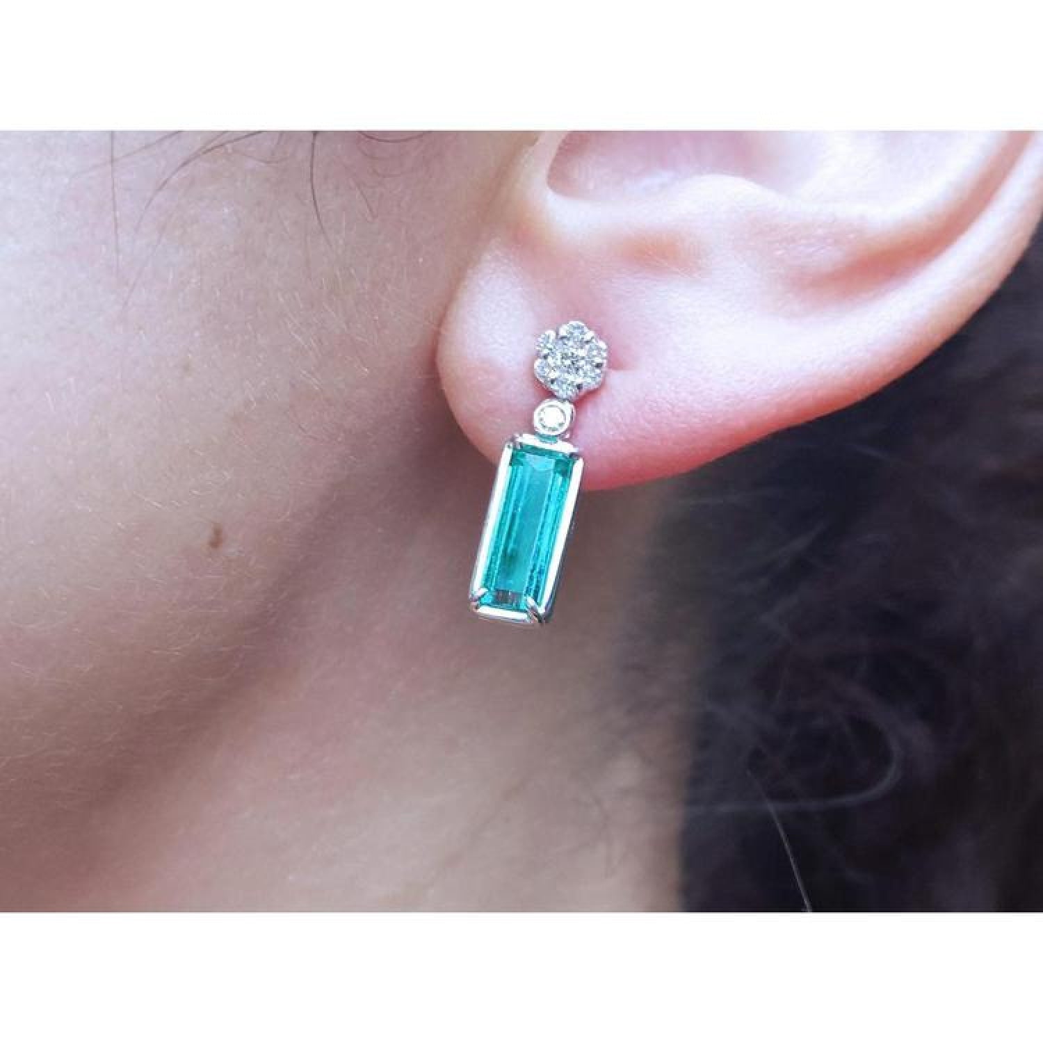 14K white gold dangling earrings with rectangular emeralds and diamond clusters on an ear.