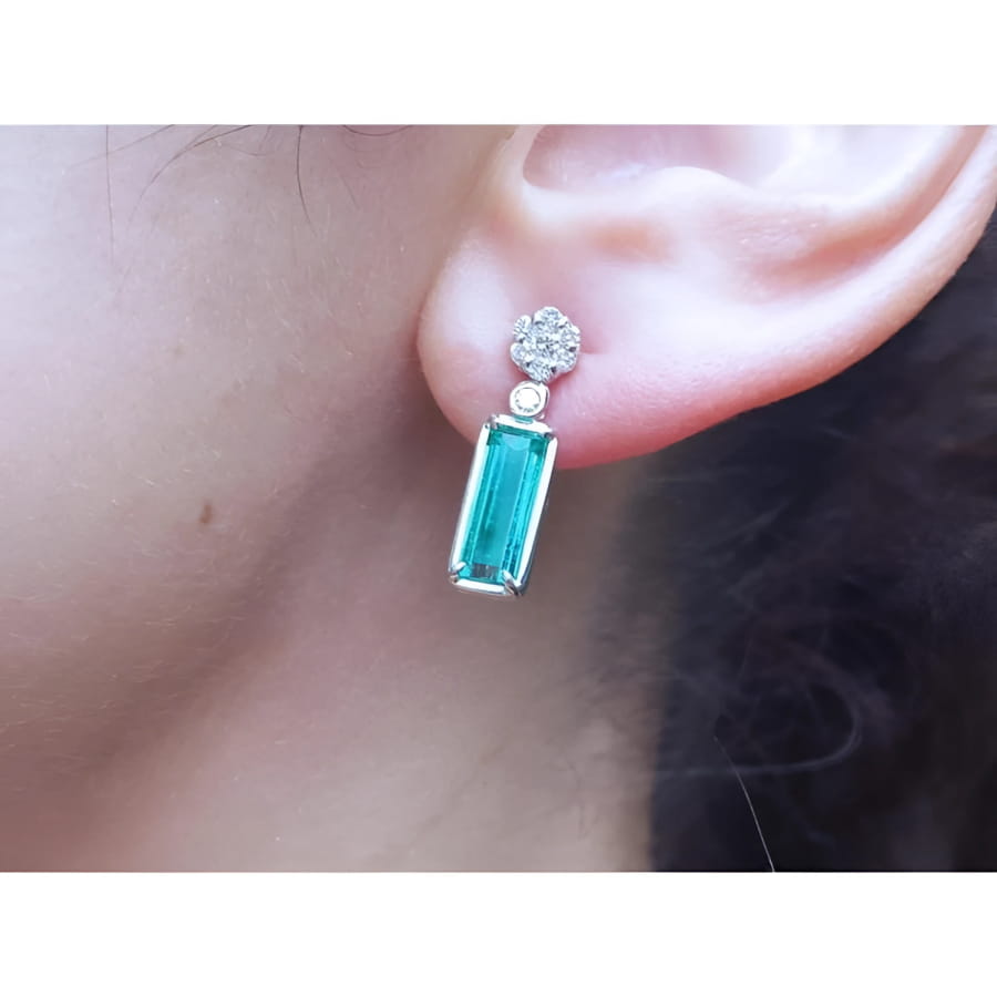 14K white gold dangling earrings with rectangular emeralds and diamond clusters on an ear.