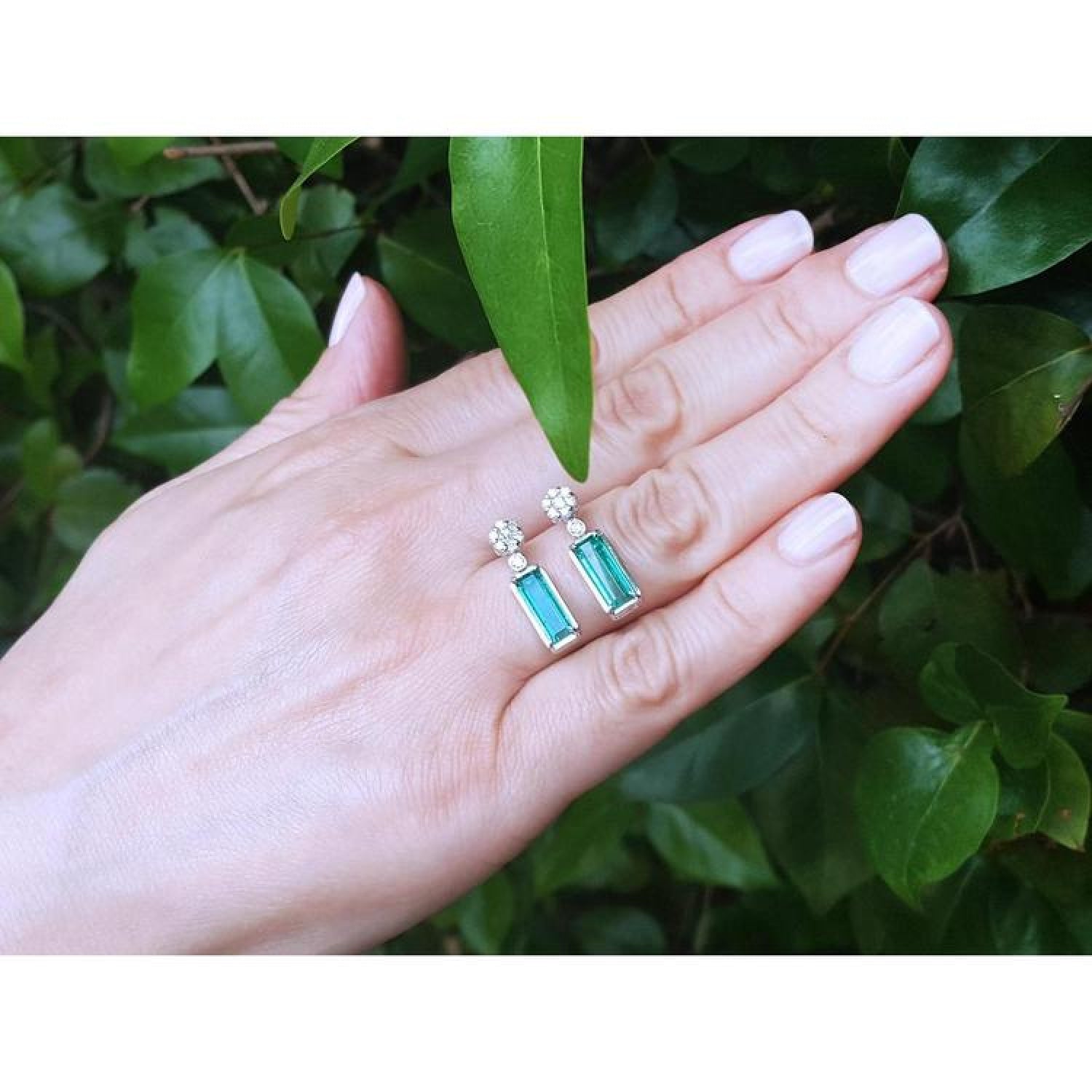 Emerald dangling earrings with diamond clusters in 14K white gold, displayed on a hand.