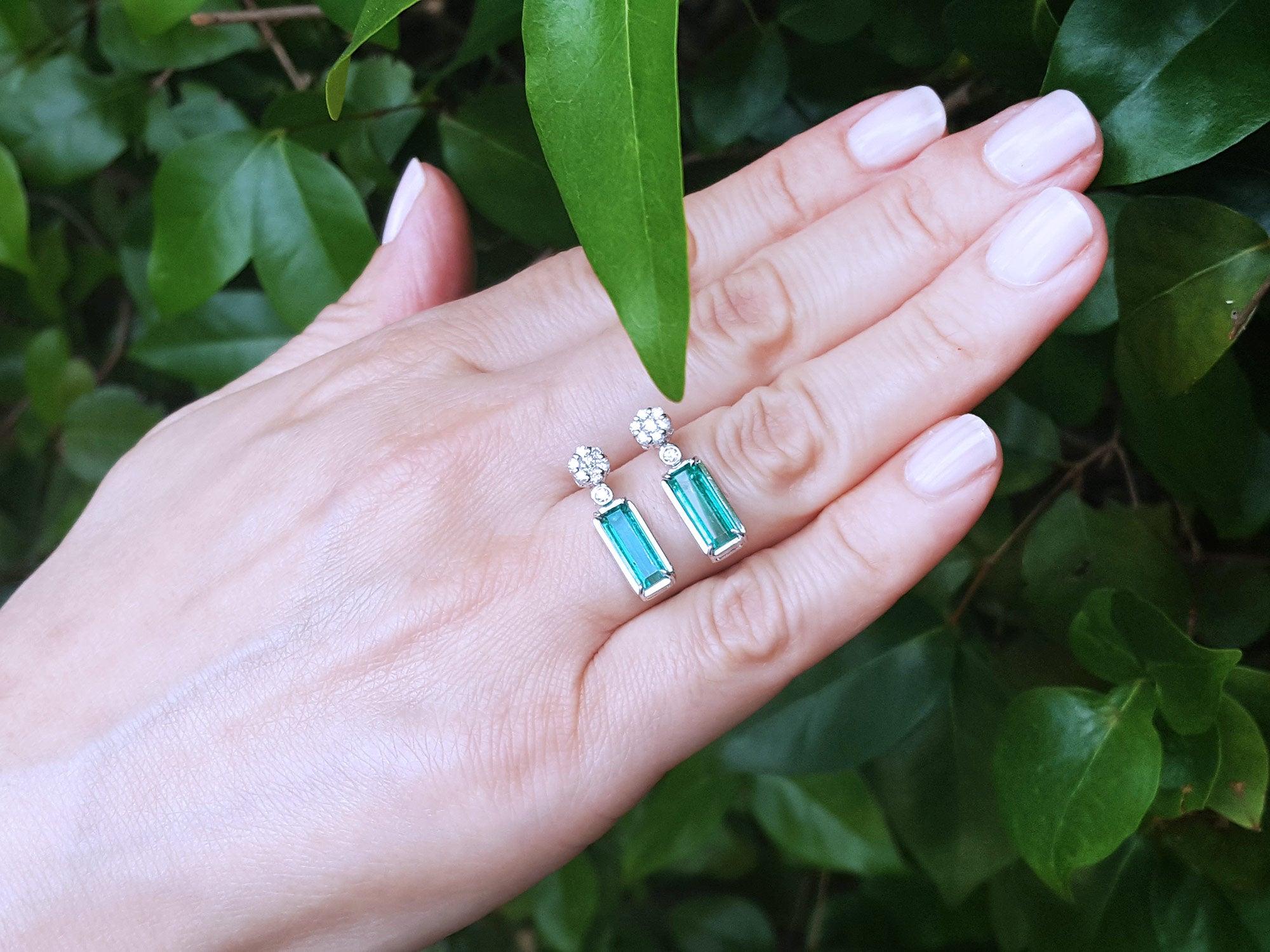 Hand displaying a pair of emerald earrings with a leafy background