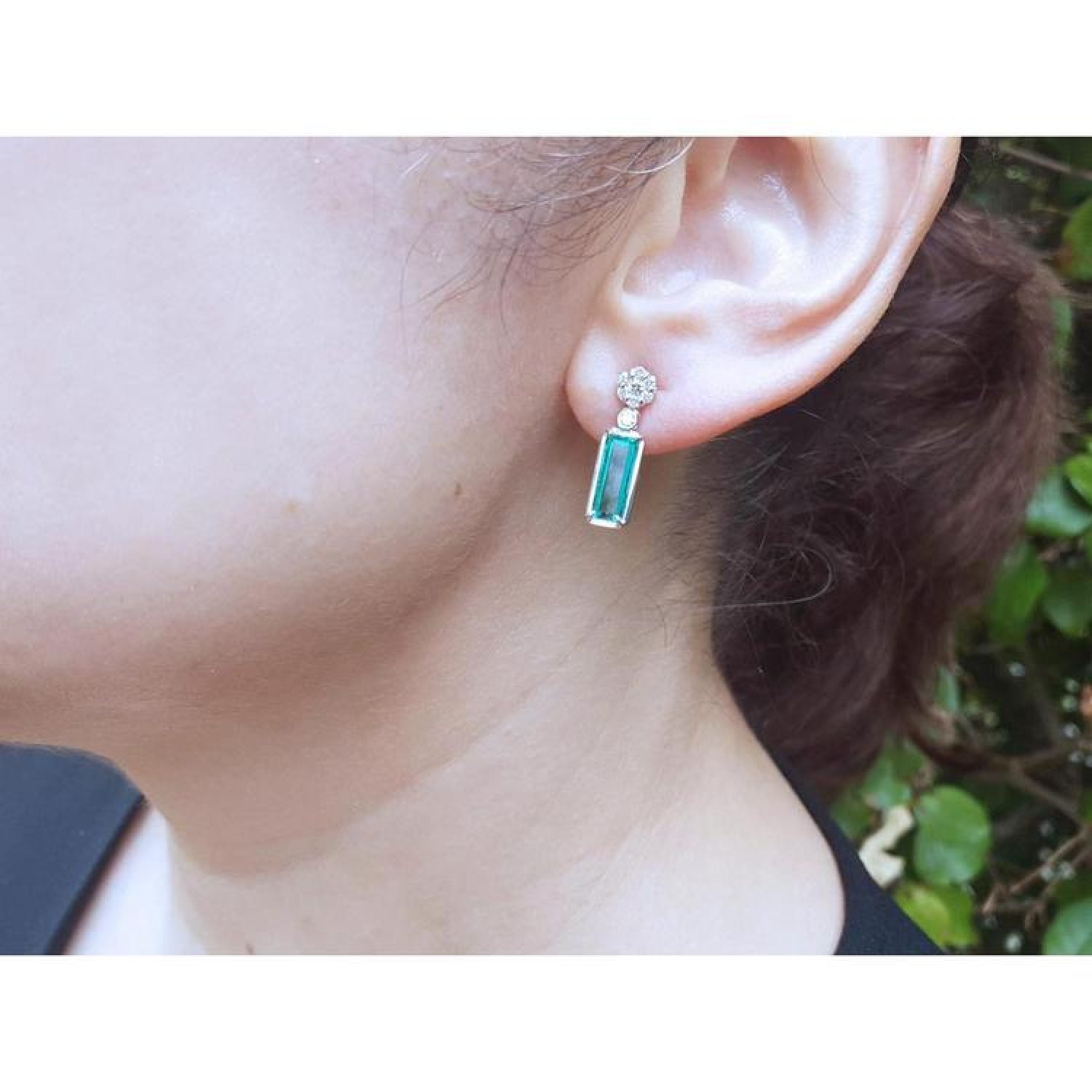 14K white gold dangling earrings with rectangular emeralds and diamond clusters on an ear.