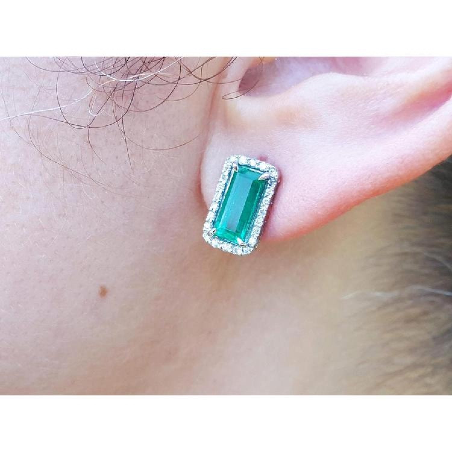 Emerald-cut Colombian emerald stud earrings with a diamond halo, set in 18K gold on an earlobe.