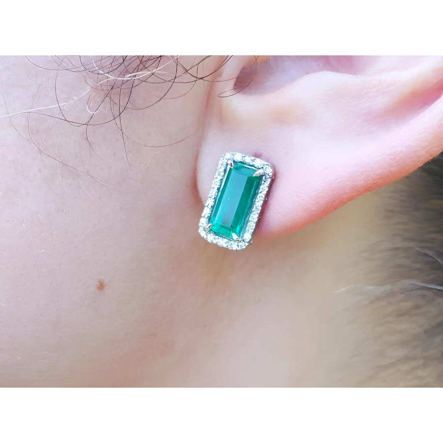 Emerald-cut Colombian emerald stud earrings with a diamond halo, set in 18K gold on an earlobe.
