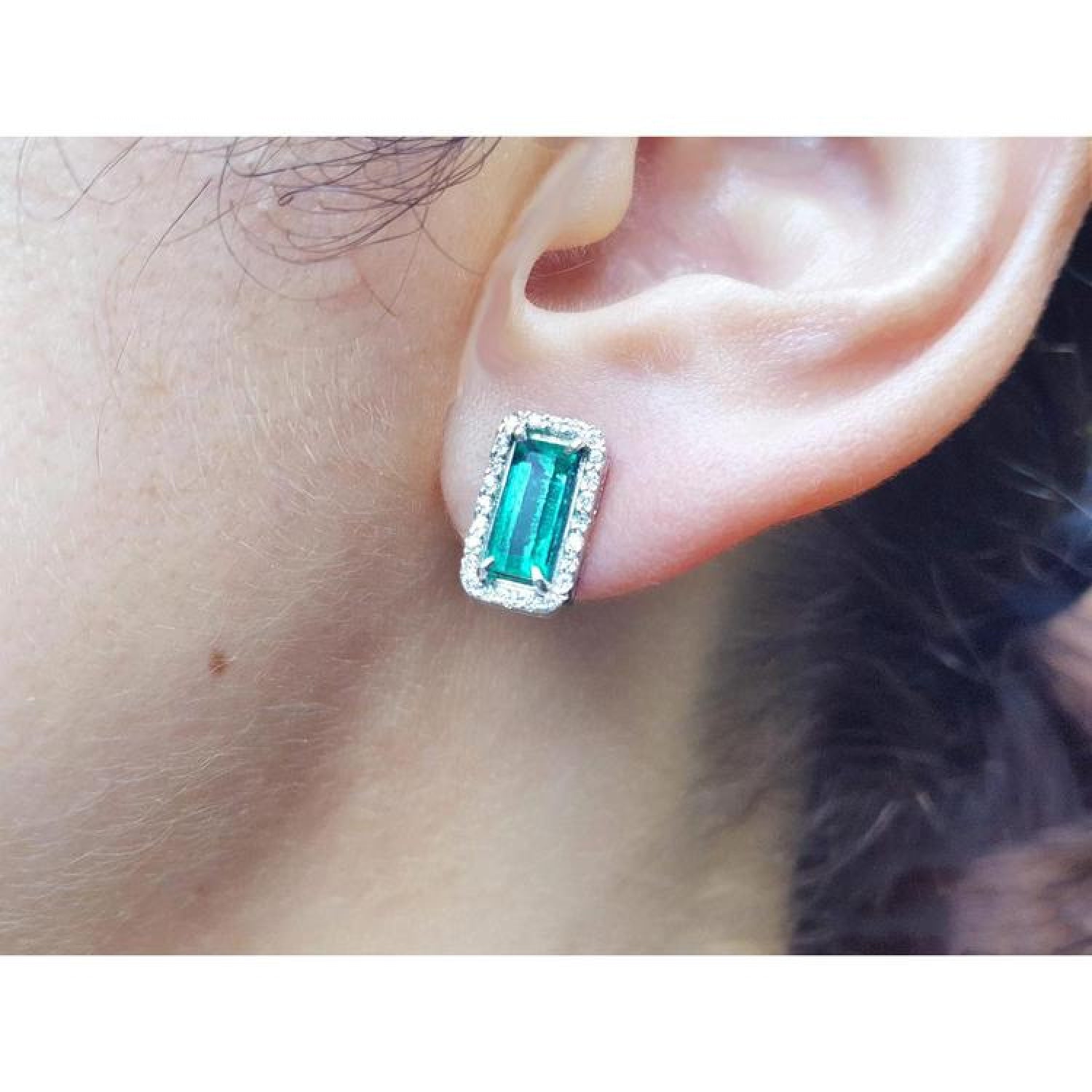 Emerald-cut Colombian emerald stud earrings with diamond halo in 18K gold, worn on an ear.