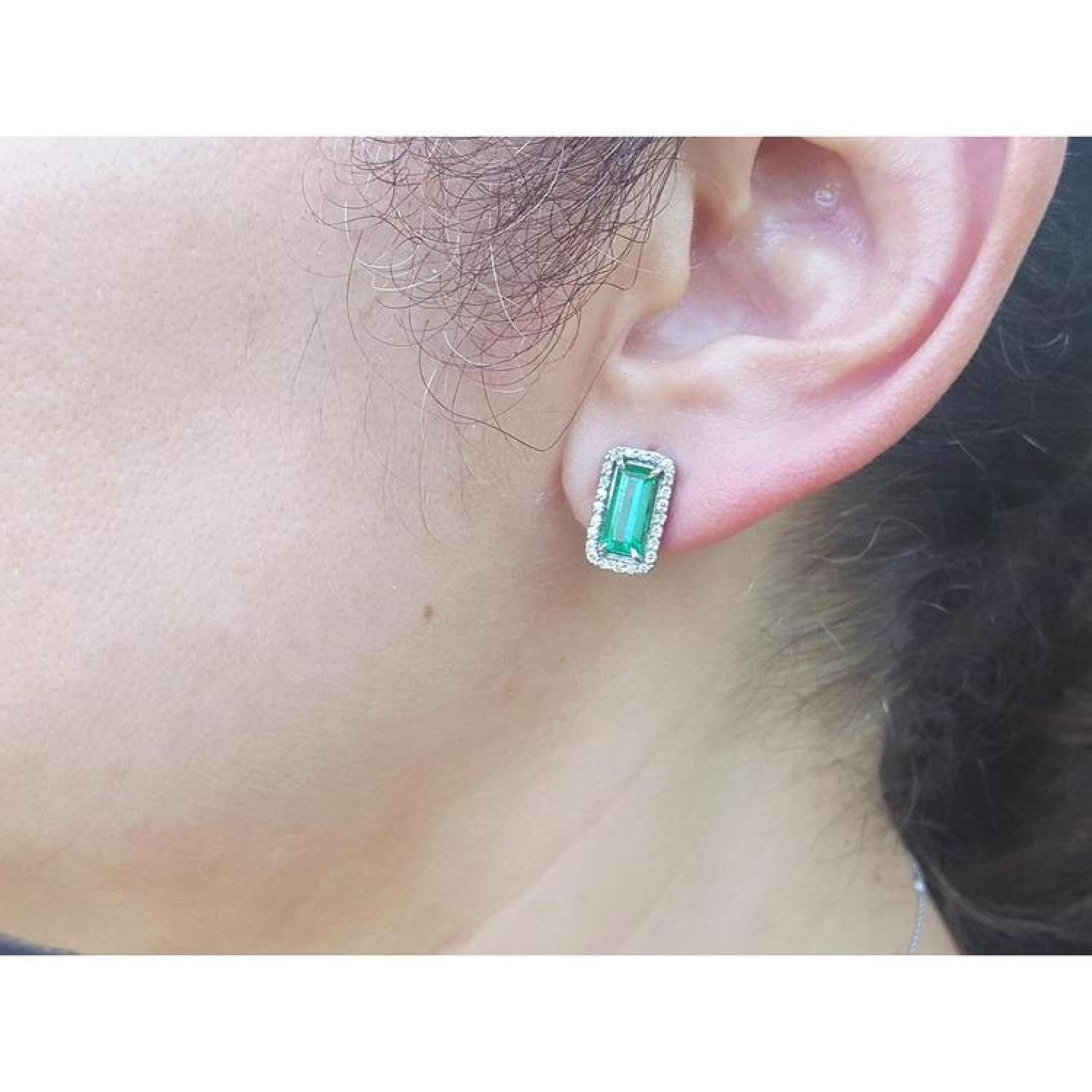 Emerald-cut emerald stud earrings with diamond halo, worn on an ear, in 18K gold.