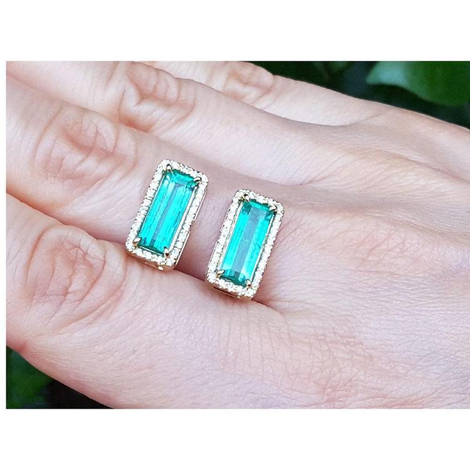 Emerald-cut Colombian emerald earrings with diamond halo in 18K yellow gold, worn on a hand.