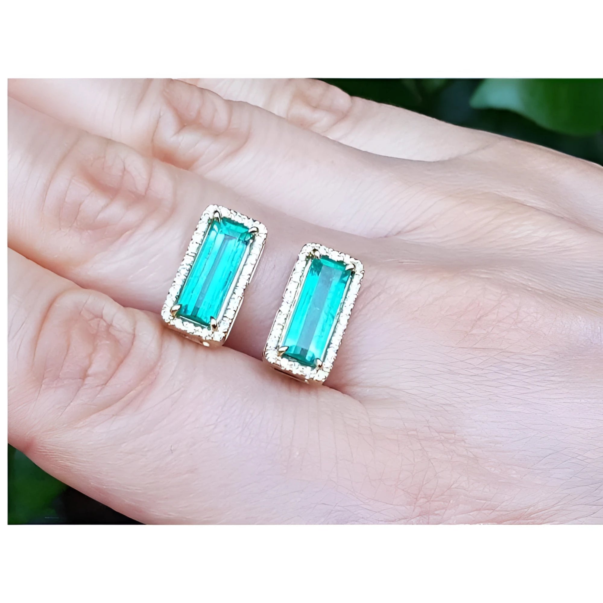 Emerald-cut Colombian emerald earrings with diamond halo in 18K yellow gold, worn on a hand.
