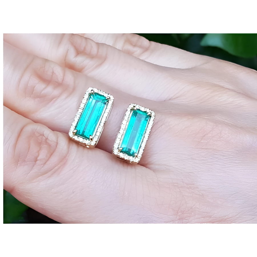 Emerald-cut Colombian emerald earrings with diamond halo in 18K yellow gold, worn on a hand.