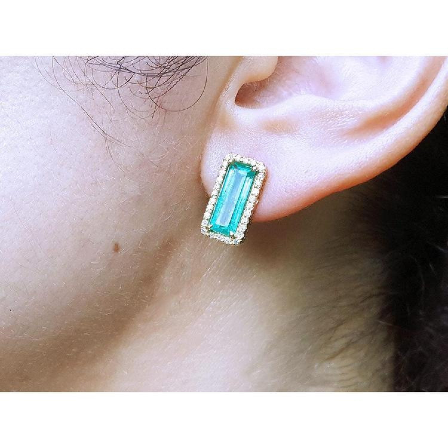 Emerald-cut Colombian emerald earrings with diamond halo, set in 18K yellow gold, worn on an ear.