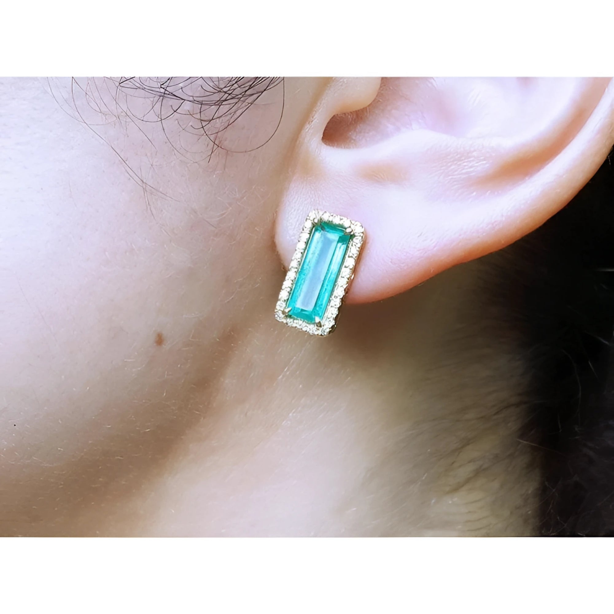 Emerald-cut Colombian emerald earrings with diamond halo, set in 18K yellow gold, worn on an ear.