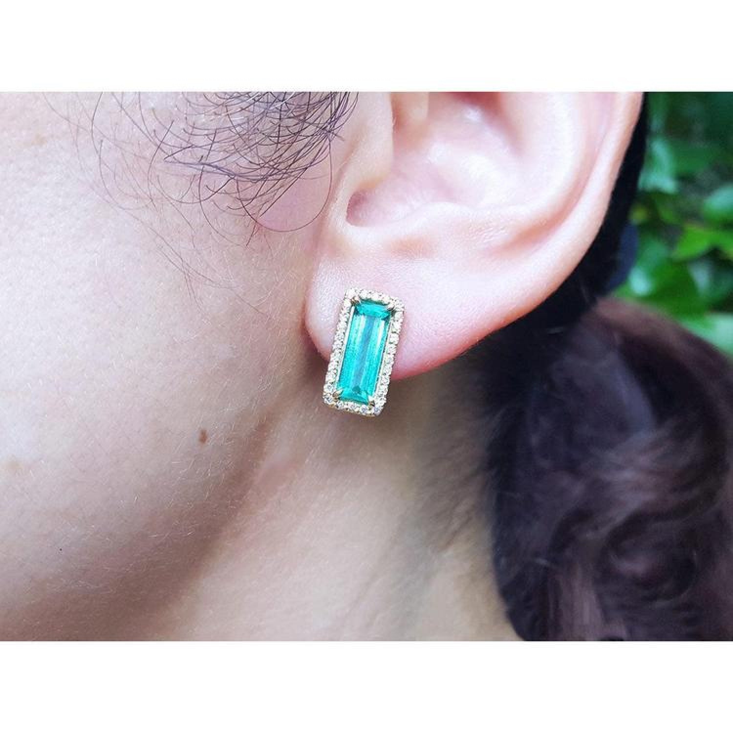 Colombian emerald-cut earrings with diamond halo in 18K yellow gold on an ear.