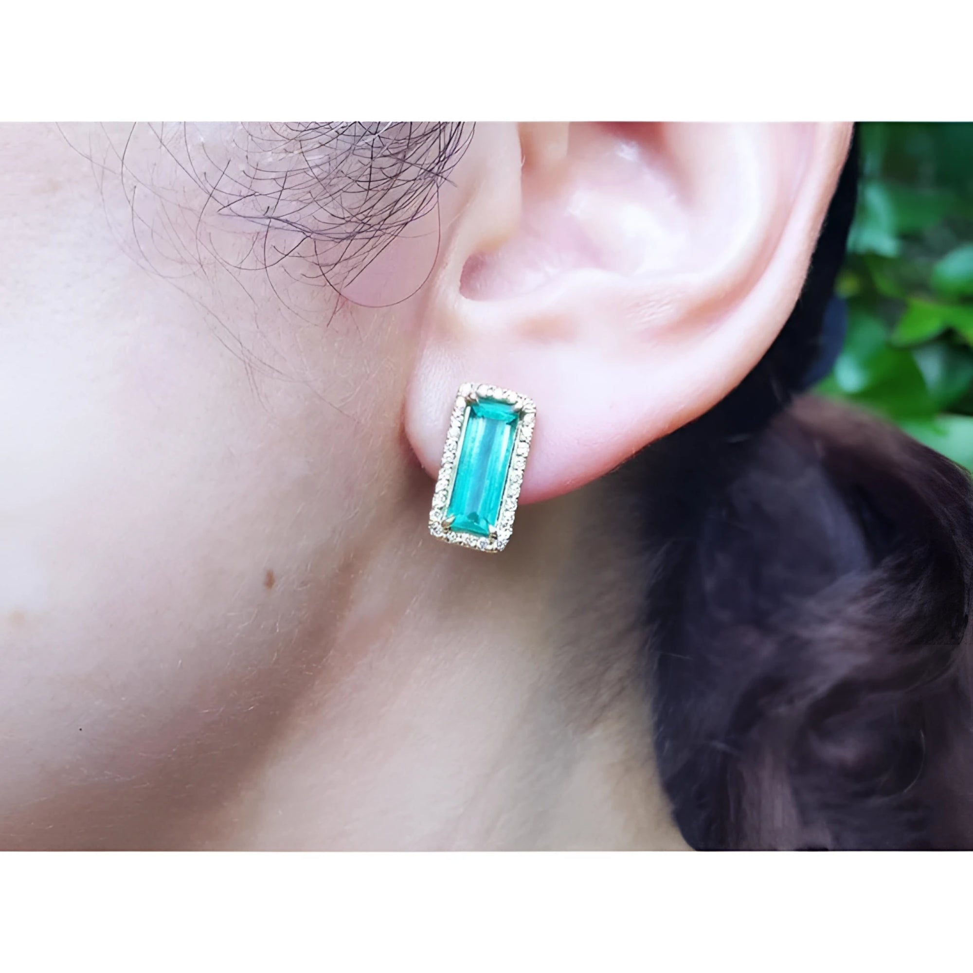 Colombian emerald-cut earrings with diamond halo in 18K yellow gold on an ear.