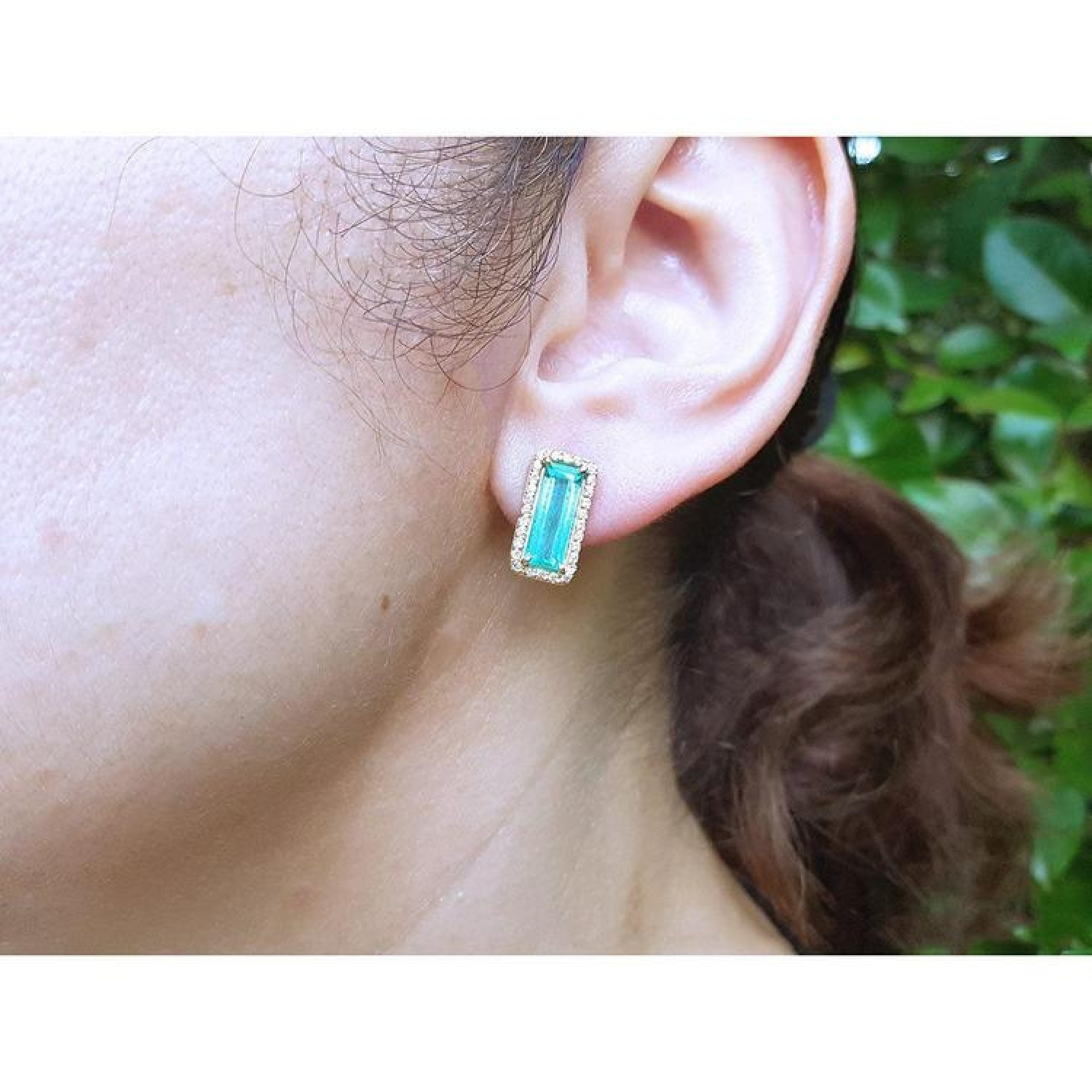Emerald-cut Colombian emerald and diamond halo earrings in 18K yellow gold on an ear.