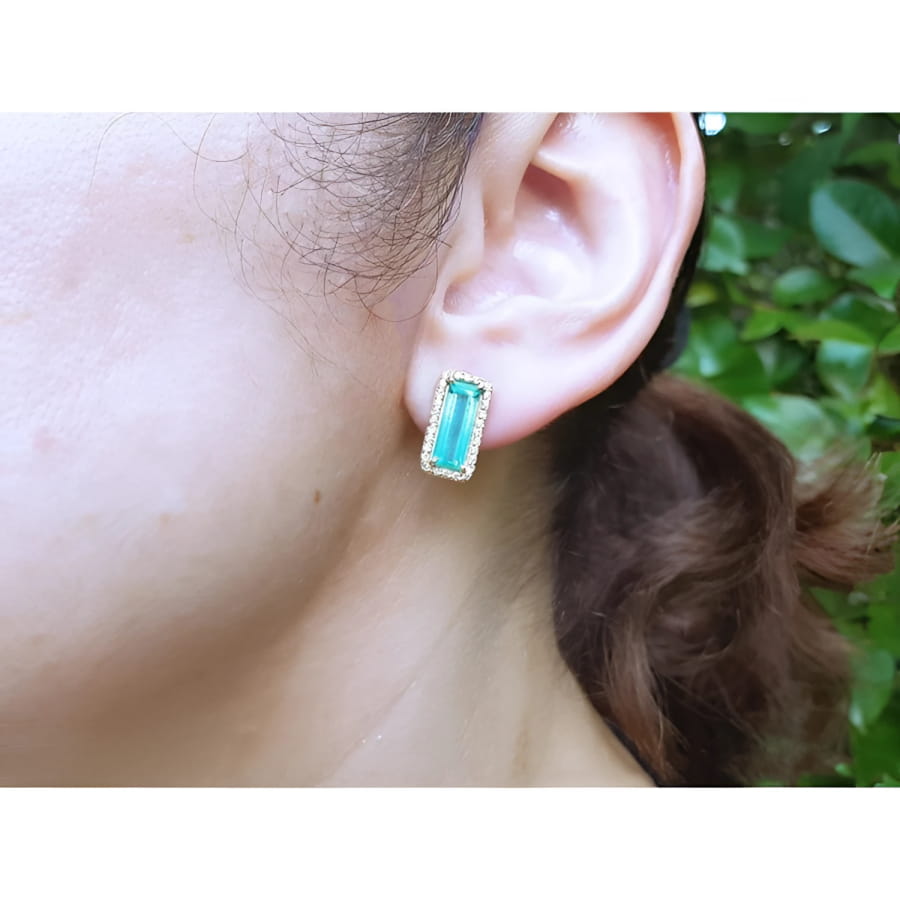 Emerald-cut Colombian emerald and diamond halo earrings in 18K yellow gold on an ear.