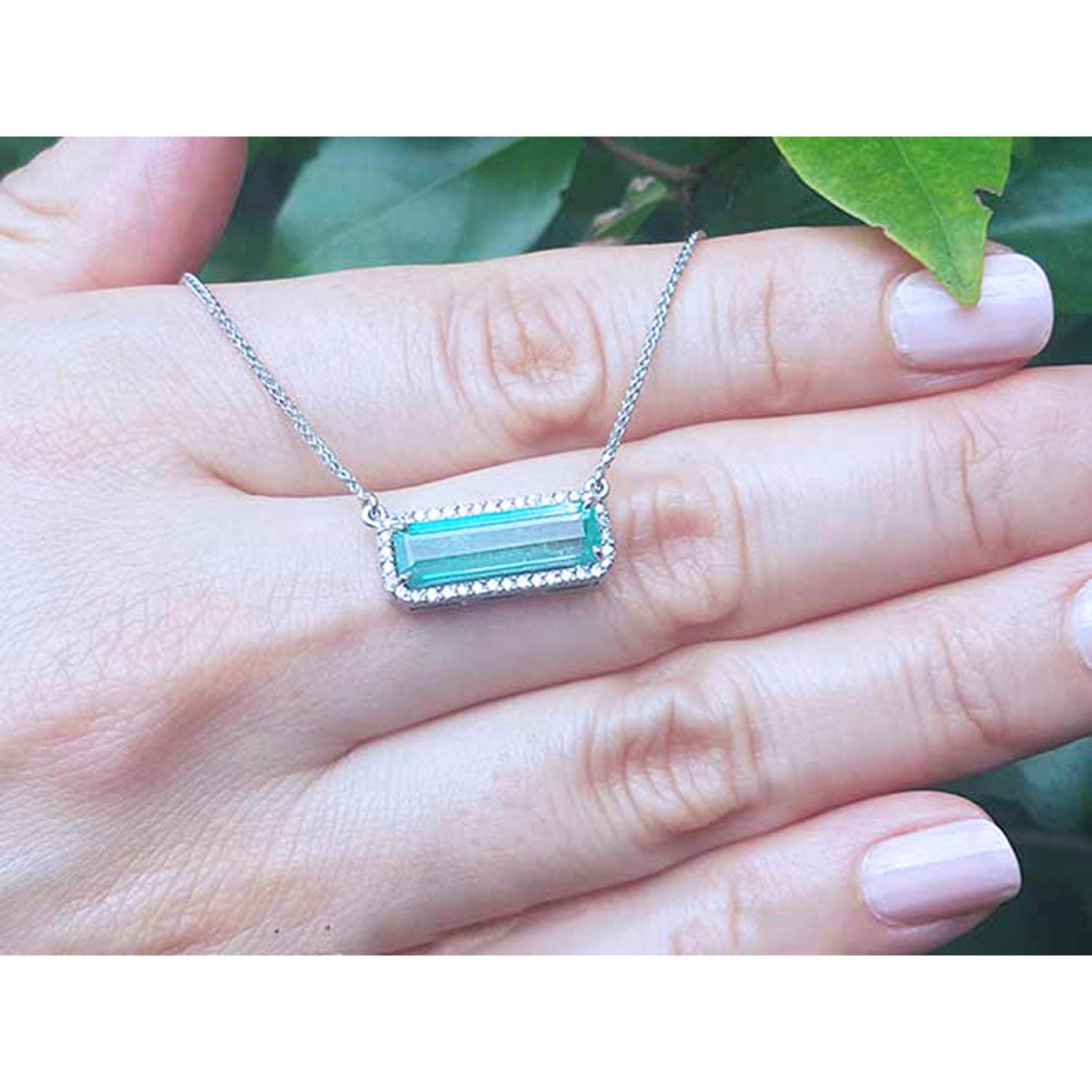 18K white gold necklace with a rectangular emerald-cut Colombian emerald and halo diamonds., held on a person's hand with green leaves in the background.