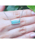 18K white gold necklace with a rectangular emerald-cut Colombian emerald and halo diamonds., held on a person's hand with green leaves in the background.